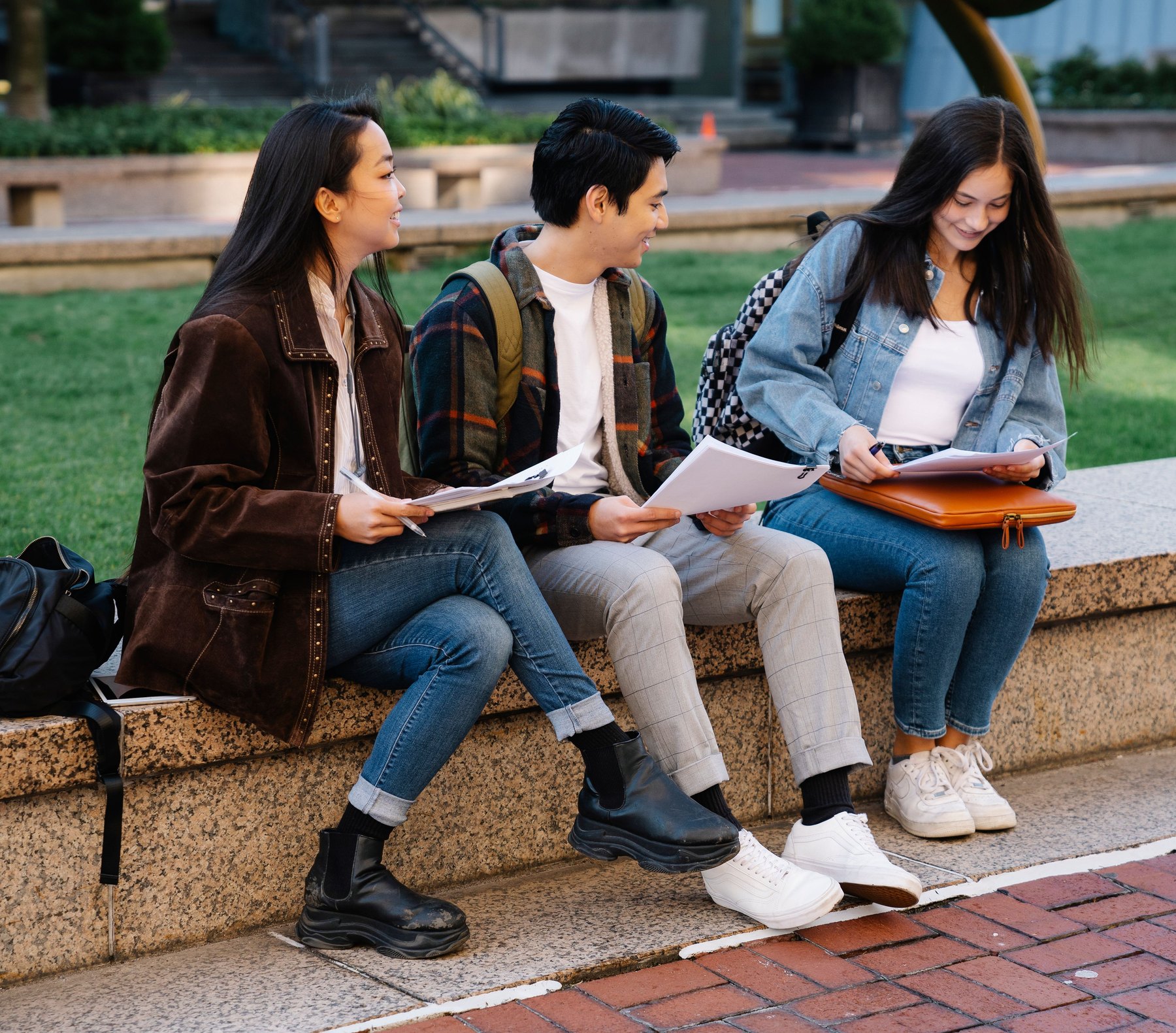 Three students sitting on a campus wall, reading paperwork together.